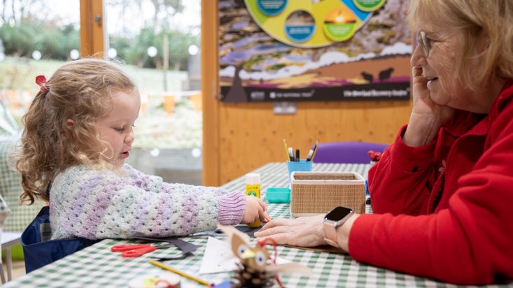 A little girl in a jumper and an older lady sat at a table covered in crafting materials.